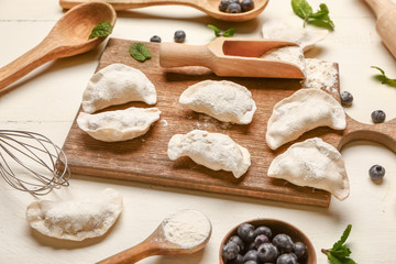 Board with raw dumplings and blueberries on white wooden background