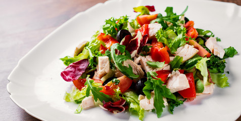 Fresh salad with chicken breast, arugula, black olives,red pepper, lettuce, fresh sald leaves and tomato on a white plate on wooden table background