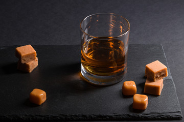 Glass of whiskey with caramel candies on a black stone slate board on black background. Side view,...