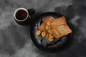 French toasts with fried plantains, dripping maple syrup, sugar powder on a black plate. French dessert concept. Sweet and delicious breakfast