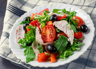 Fresh salad with chicken breast, arugula, black olives,red pepper, lettuce, fresh sald leaves and tomato on a white plate on wooden table background