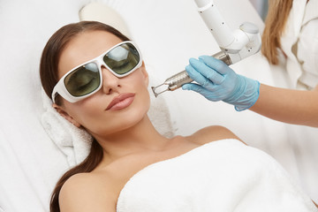 beautician wearing gloves and holding laser near beautiful woman lying with protection glasses in beauty salon