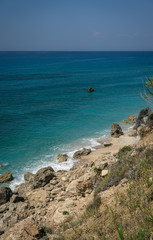 Beautiful rocky seashore, sunny beach with turquoise water. Stone, sand, waves and plants. Blue and green, bright. No clouds sky.