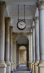 A large old vintage clock hanging from the ceiling and a view of a passageway with columns
