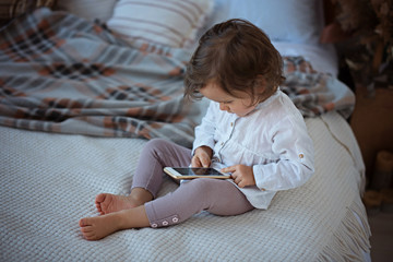 Little girl sitting on the bed holding a smartphone, playing games and watching cartoons. Baby addiction to gadgets.
