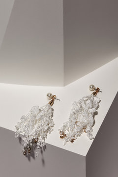 Subject shot of a pair of earrings isolated on the white surface in the space with geometric design. Each earring is made as a pearl with white lace pendants with sparkling crystals in golden setting.
