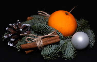 Art installation on a black background for New Year or Christmas from spruce twigs, one glass ball,  pine cone, one mandarin and cinnamon sticks.