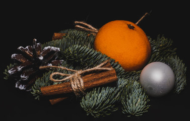 Art installation on a black background for New Year or Christmas from spruce twigs, one glass ball,  pine cone, one mandarin and cinnamon sticks.