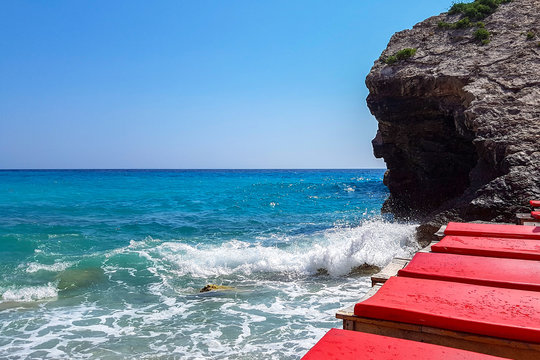 Panoramic view of pebble beach with clear azure blue water, layered rocks and red beach umbrellas, beautiful mediterranean Adriatic Sea coast, Montenegro, selective focus