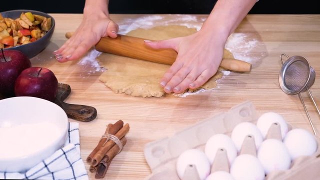 Female hands rolling a dough with rolling pin on the table with different baking ingredients close-up.