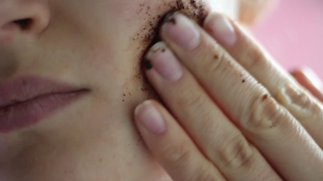 Girl puts coffee scrub on her face close-up pink background.