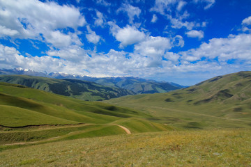 Fototapeta premium Road over green hills, mountains in snow and clouds in blue sky