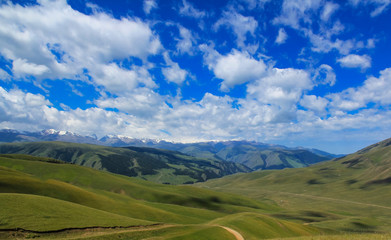 Road over green hills, mountains in snow and clouds in blue sky