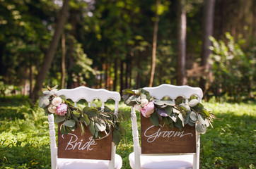 Luxury decorations for wedding day. White chairs with wooden plaques on which inscription of bride and groom are decorated with fresh eucalyptus leaves, pink peonies and white roses. Green Forest.