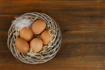 Raw chicken eggs and feather in wicker nest on wooden table, top view. Space for text
