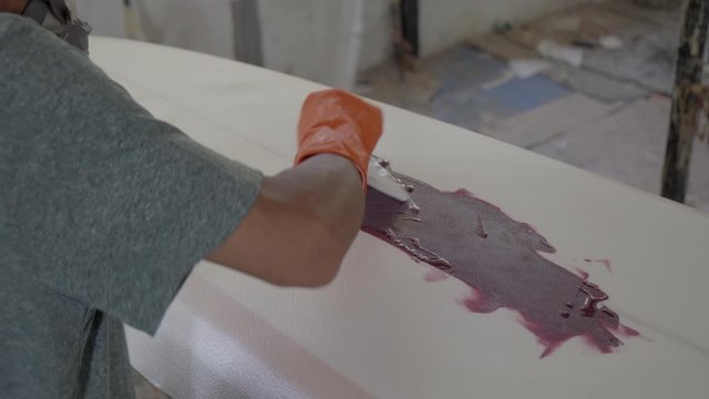 Sequence of shots of Indonesian worker wearing safety mask painting fabric covered surfboard using special tool