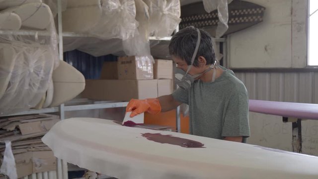 Sequence of shots of male Indonesian worker wearing safety mask and rubber gloves painting fabric covered surfboard with special tool