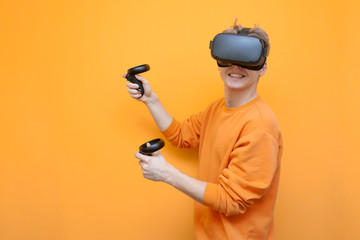 joyful guy in VR glasses on an orange background, a gamer plays a virtual game and holds modern joysticks