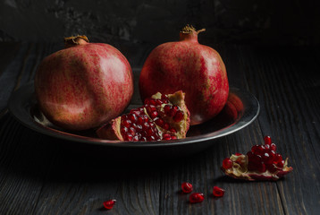 Two pomegranates lie on a metal plate, which stands on a dark wooden table. About them is part of the third broken grenade. On the table lies a piece of pomegranate. The dark key.
