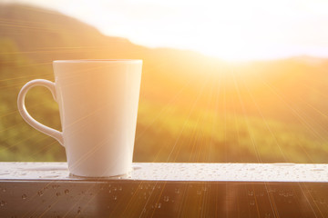 A white cup of coffee is on a balcony in the morning after rained, with mountain view and light of...