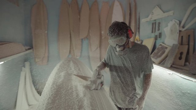 Sequence of shots of Indonesian craftsman wearing protective mask, goggles and headphones sanding surfboard using electric sanding machine