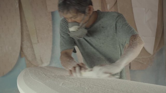 Sequence of shots of Indonesian surfboard shaper wearing protective mask sanding surfboard with abrasive paper
