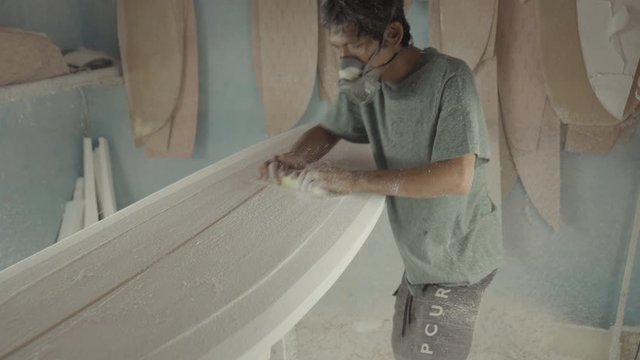 Sequence of shots of Asian surfboard shaper wearing protective equipment sanding new handmade surfboard lying on workbench using emery paper
