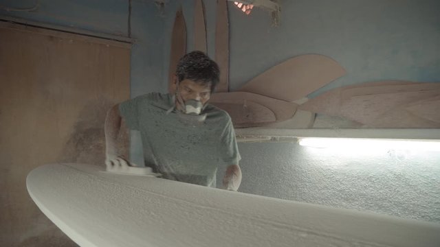Sequence of shots of professional surfboard shaper wearing protective mask polishing surfboard using abrasive paper