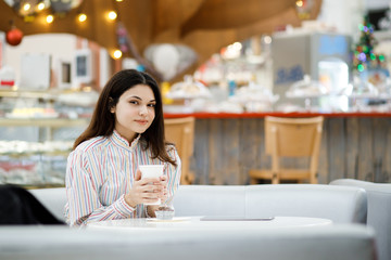 Pretty young brunette girl sitting in cafe and holding tablet, drinking coffee and resting