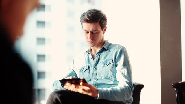 Thoughtful young business man is using tablet in the office against the background of a window and glass buildings. Camera's coming from behind man's back. Tracking shot in slow motion.