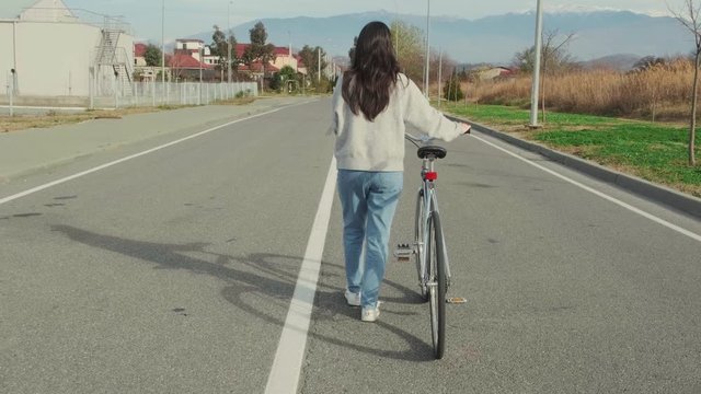A young brunette woman in casual clothes is walking down the center of the road with a retro bike.