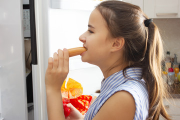 The girl looks in the refrigerator, hungry, eats sausage