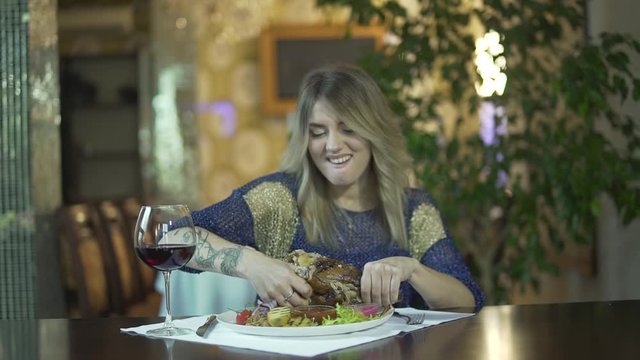 Young tattooed beautiful blonde girl in fancy dinner restaurant tearing meat eating with fingers inappropriate behaviour