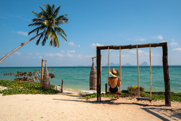 Isolated woman on her romantic honeymoon getaway on a paradisiacal beach with palm trees. Expensive luxury resort for married couples and single people. Relax on a swing. Travel to Thailand concept.