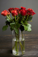 Bouquet of red-orange roses in a glass vase on a dark wooden surface, greeting background or concept