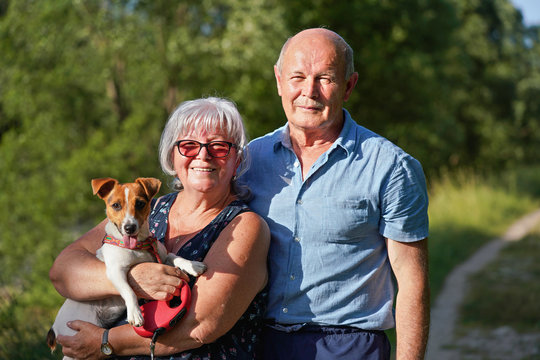 Couple of seniors posing with their small Jack Russell dog, tongue out. Woman holds her on hands, blurred trees and country road background