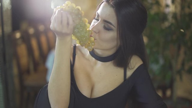 Close up shot of seductive sexy young brunet woman in black cleavage dress eating grape fruits alone at fancy restaurant
