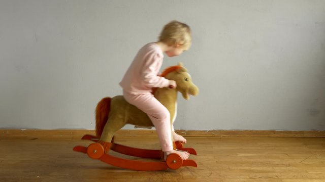Funny blond boy in pink pajamas riding horse on rocking chair against white wall