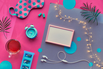 Healthy night sleep creative flat lay. Frame with copy-space. Sleeping mask, blue mint alarm clock, earphones, earplugs and tea. Split pink and silver background with circles and palm leaves
