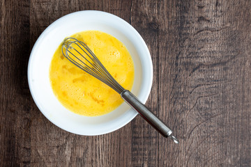 Raw whisked eggs in white bowl with metal whisk, wood table