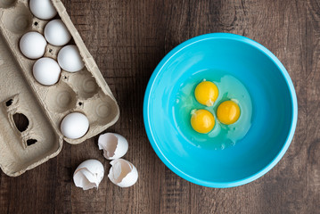 Fototapeta na wymiar Three cracked eggs in blue mixing bowl, eggshells, whole eggs in cardboard carton, wood table