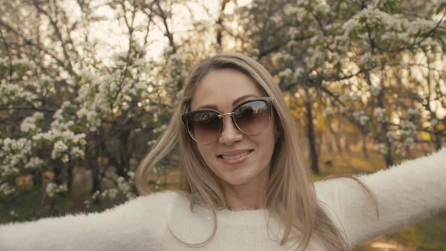 Attractive young woman in sweater, jeans is dance and spinning among blossom apple tree. Sunset, spring orchard and green field. Slow motion. Caucasian one girl, long fluttering hair. Sun glasses.