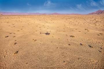 An aerial view of a Namibia landscape, with the shadow of a tourist sightseeing helicopter visible on the sandy ground.