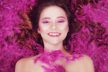 Happy brunette is smiling. Girl lezhin on a background of purple feathers, pink background. Girl with a beautiful roam