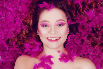 Happy brunette is smiling. Girl llies on a background of purple feathers, pink background. Girl with a beautiful roam / purple make-up.