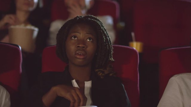 Tracking shot of black woman watching thrilling movie at cinema. She is looking at screen with her eyes wide open and eating popcorn