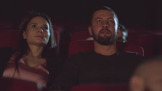 PAN shot of man and woman watching horror movie in theater and getting scared