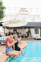 Pretty young Asian women sitting on edge of swimming pool, drinking wine and discussing new mobile appication
