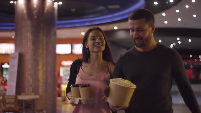 Tracking shot of happy young woman with two cups of coffee and bearded man with bucket of popcorn walking and chatting at cinema