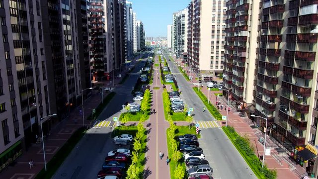 Aerial view of beautiful city street under the suumer sunshine with many walking people and driving cars. Motion. Flying above the houses, buildings, green lawn along the road.